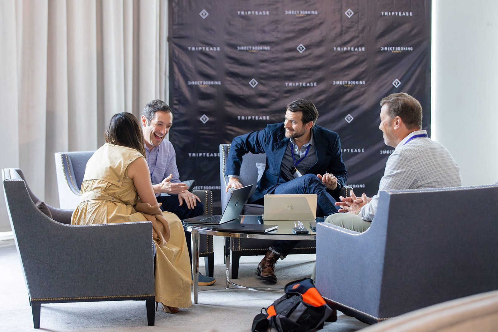 Four attendees in armchairs sharing a laugh in front of a Triptease Direct Booking Summit backdrop